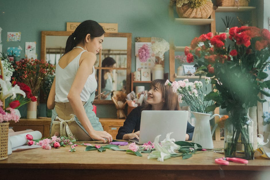 A florist and a customer engaged in a conversation inside a flower shop. The florist, wearing a white sleeveless top and beige apron, stands beside a wooden table covered with various pink and white f