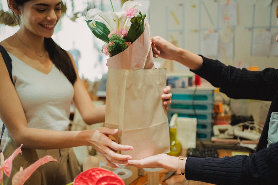 A smiling female florist with long dark hair wearing a beige apron and white top, is seen handing over a bouquet of fresh flowers inside a brown paper wrapping to a customer, whose arm is visible reac