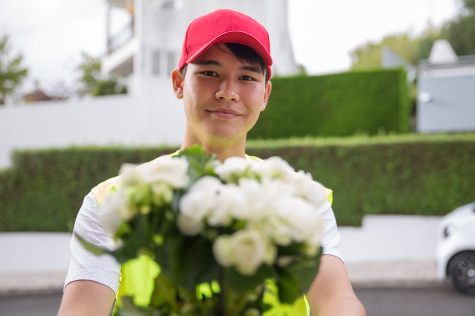 A delivery person wearing a red cap and high-visibility vest is smiling and extending a bouquet of fresh white flowers towards the camera. The arrangement features tightly packed white blooms with gre
