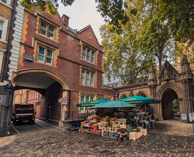 A vibrant flower stall situated on a cobblestone street near a historic stone archway and red-brick building with arched windows. The stall, operated by flowerdeliverydenmarkhill.co.uk, displays a col